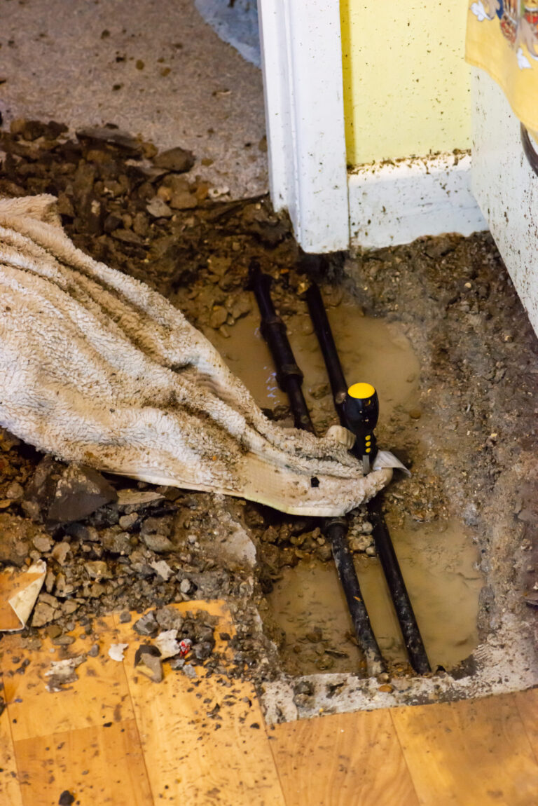 Exposed pipes run through an opened concrete floor with standing water, rubble, a towel, and a hand tool.